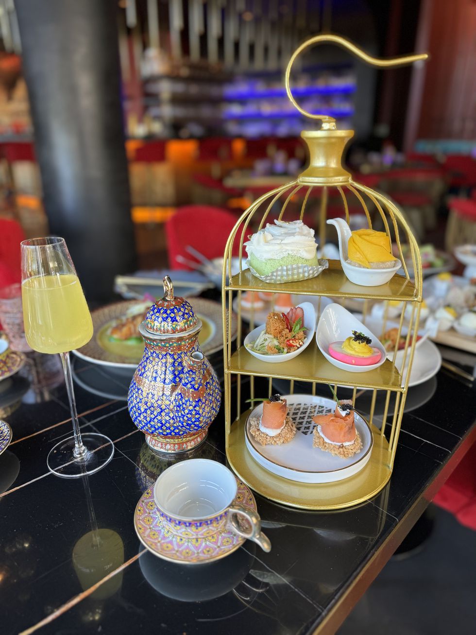 A high tea spread that includes a Thai tea kettle and cup, and a tiered circular tray with sandwiches and sweets.