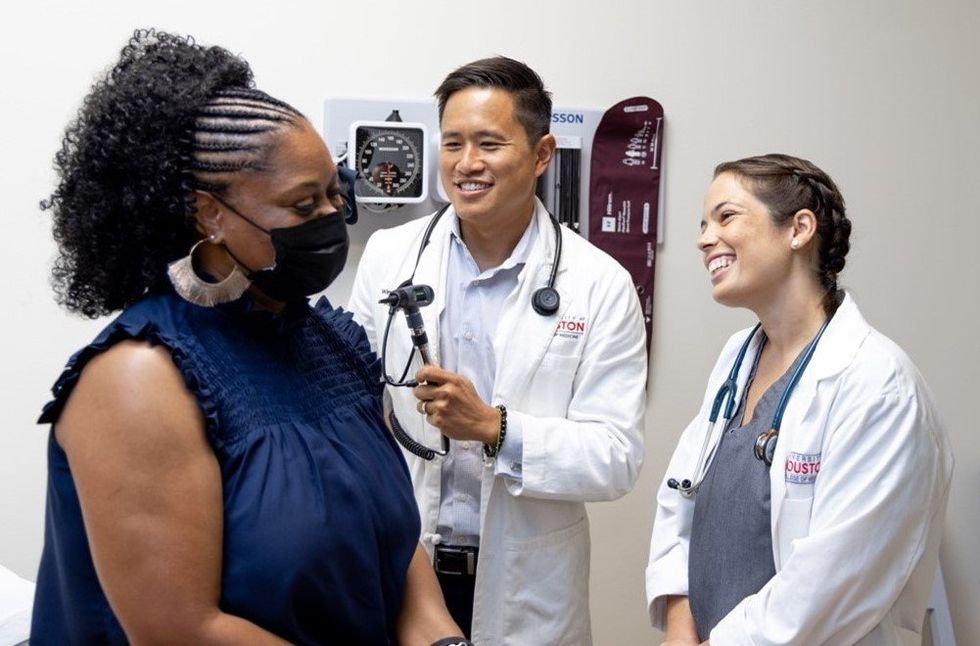A man and woman in lab coats talk to a female patient in a blue shirt wearing a mask. They are in an examining room.