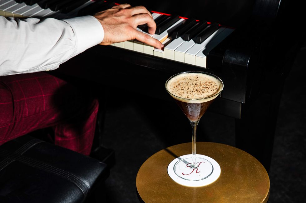 A man plays the piano while a cocktail sits next to him