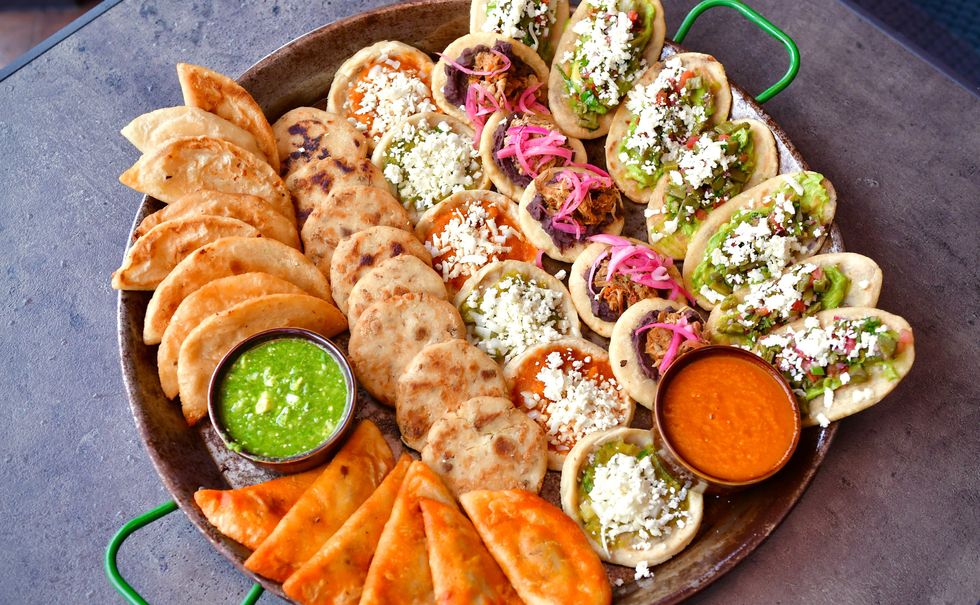 A round platter with green handles holding red and green salsas, empanadas and soft tacos filled with meats and cheeses.