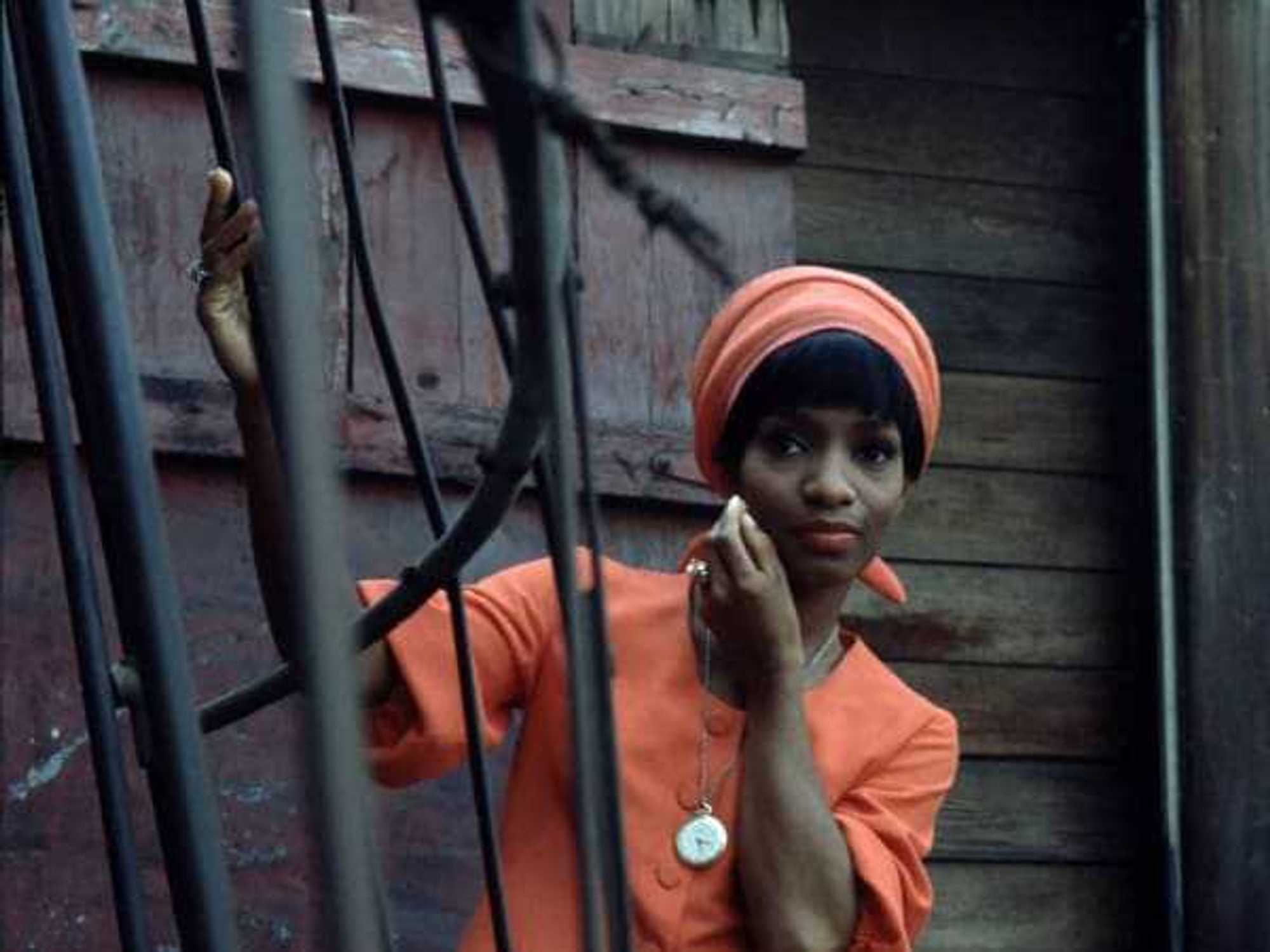 Color photo by LaMonte McLemore of a Black woman in a bright coral suit set against dark brown wood and a black iron fence