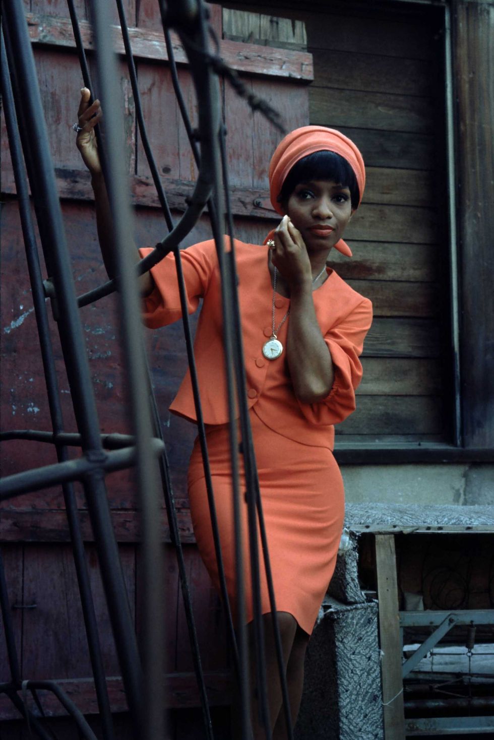 Color photo by \u200bLaMonte McLemore of a Black woman in a bright coral suit set against dark brown wood and a black iron fence