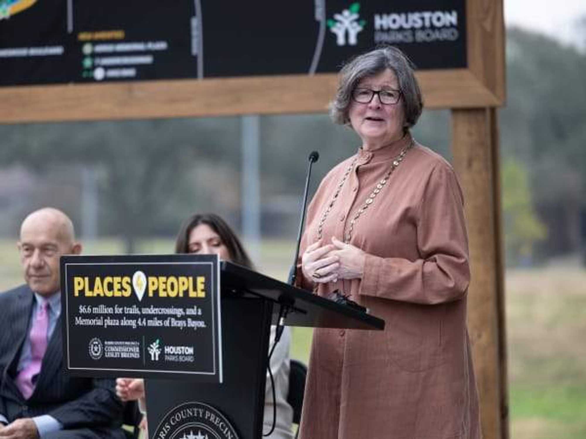 Houston Parks Board CEO Beth White stands at a podium at the Captain Herod Memorial, Houston Mayor John Whitmire is seated nearby.