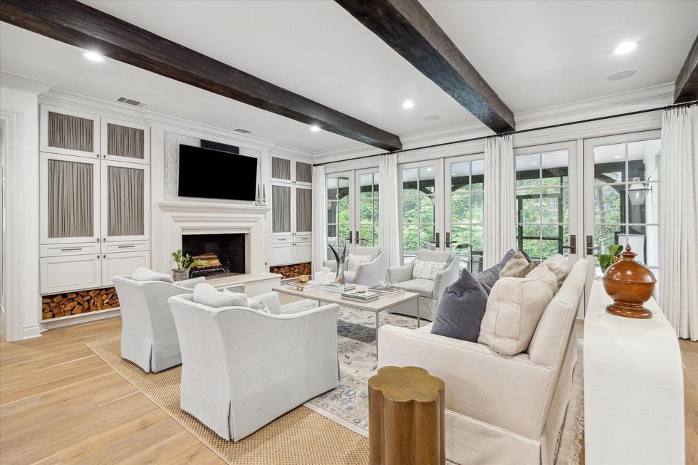 Image of a big living room with hardwood floors. Two chairs are in the foreground, two more in the background. A sofa with pillows is between them, forming a U and looking toward a fireplace. A flatscreen TV is above the fireplace.
