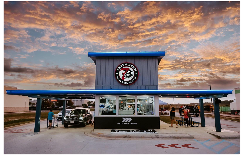 Image of a drive-through building with cars at the window, and a small seating area on the right side. The round 7 Brew logo is above the drive through roof.