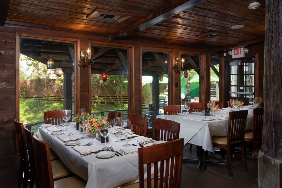 Image of the dining room at Rainbow Lodge, with a table for six in the foreground, with a white tablecloth and a vase of orange lilies,; the room overlooks the wooded bayou.