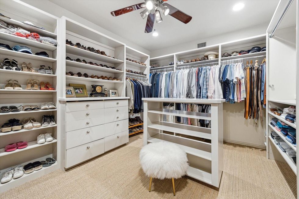 Photo of a closet with a shelf of shoes, a built-in dresser, an island shelf unit, a big poofy stool and having racks on three sides.