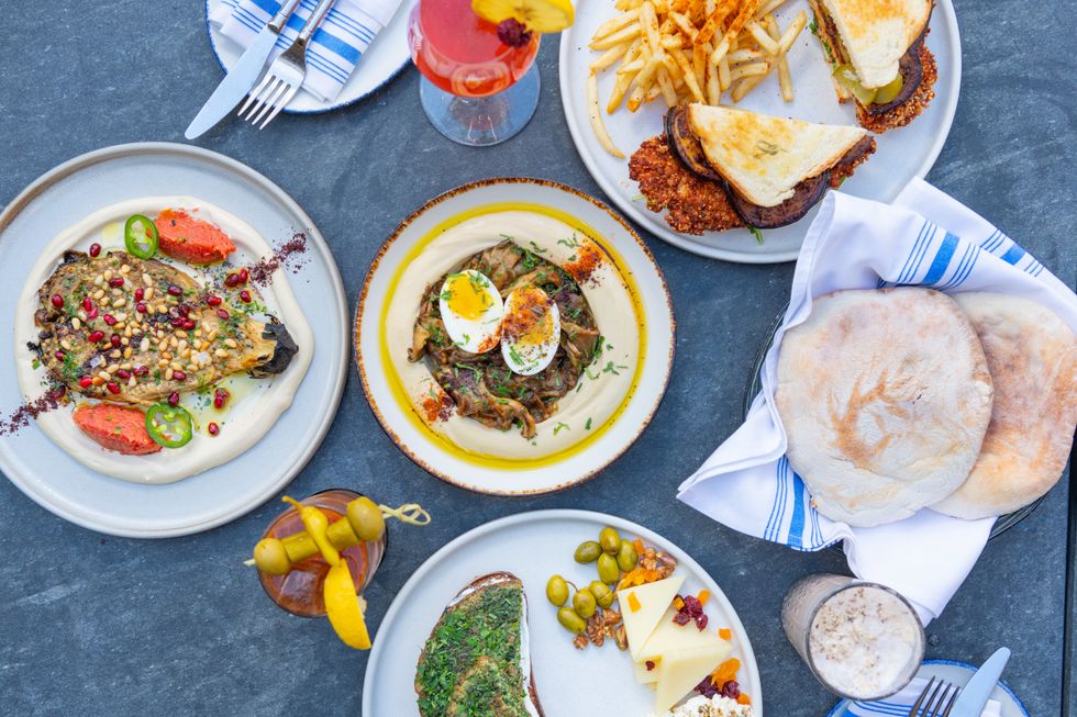 Photo of a collection of white plates with brunch items on them, including pita bread, hummus and lamb and a Bloody Mary.