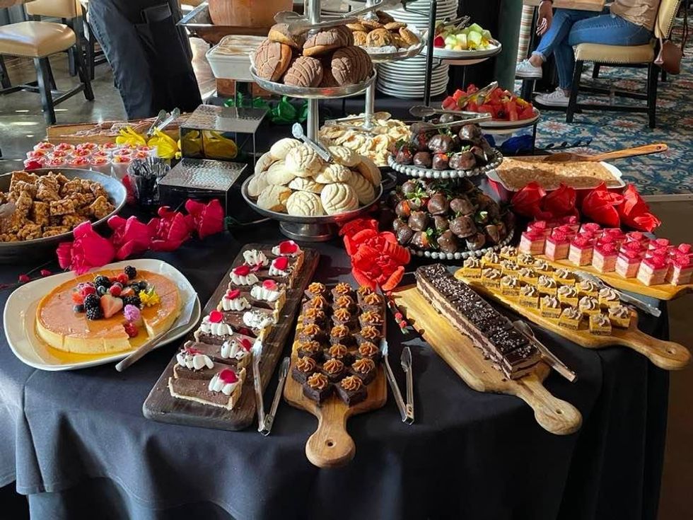Photo of a table with many dessert platters on it, with items such as cheesecake, brownie bites, cookies and fruit.