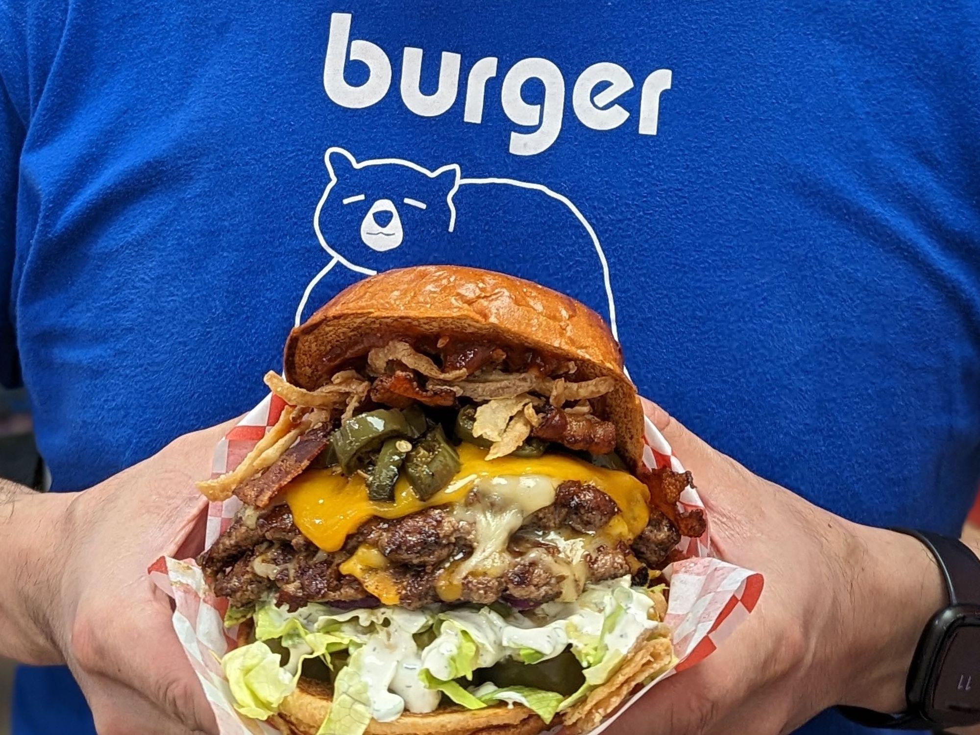 Photo of chef Willet Feng holding a double burger with lettuce, cheese, jalapenos, bacon and onion strings.