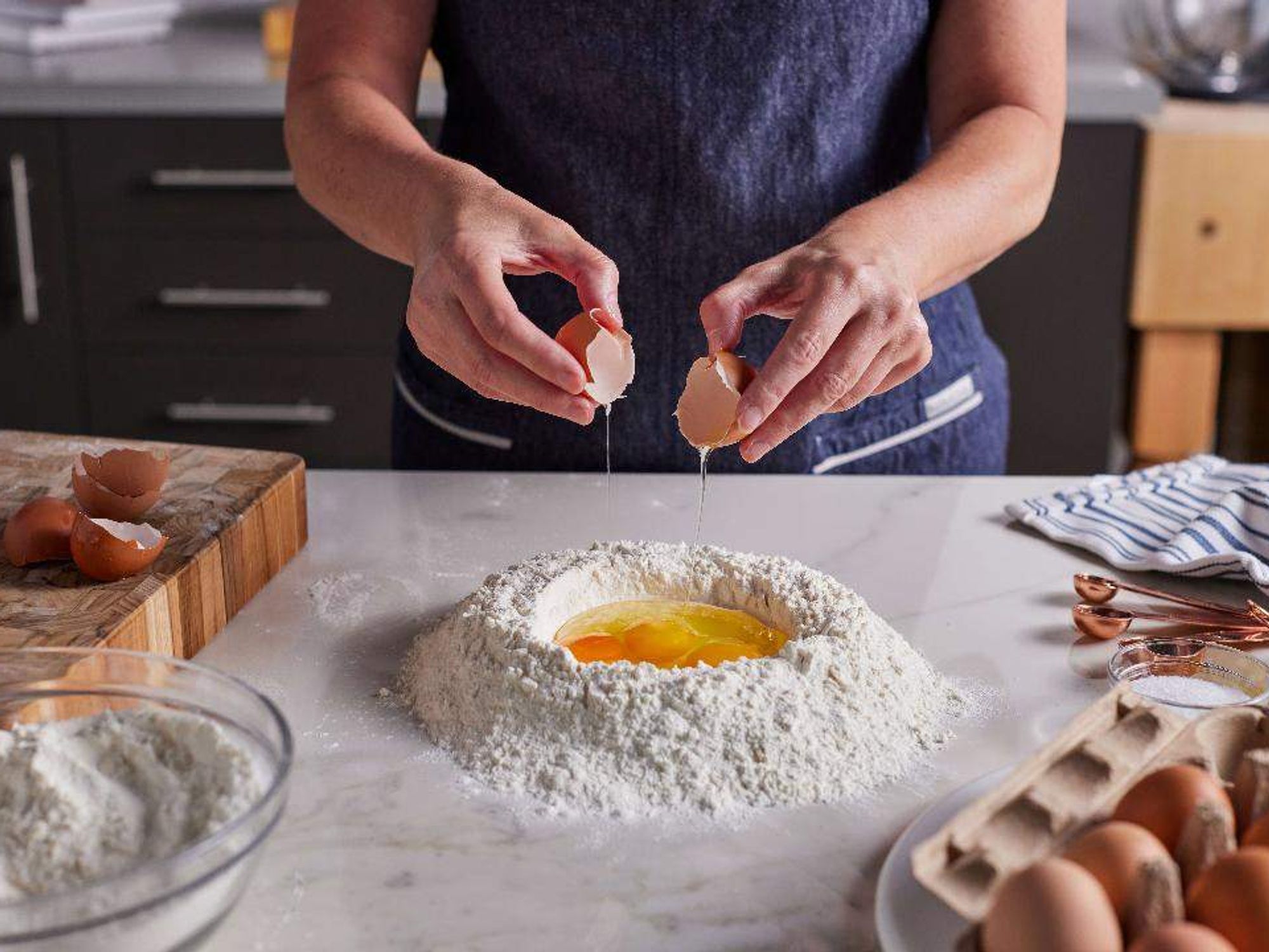 Picture of a pair of woman's hands cracking an egg into flour on a marble counter