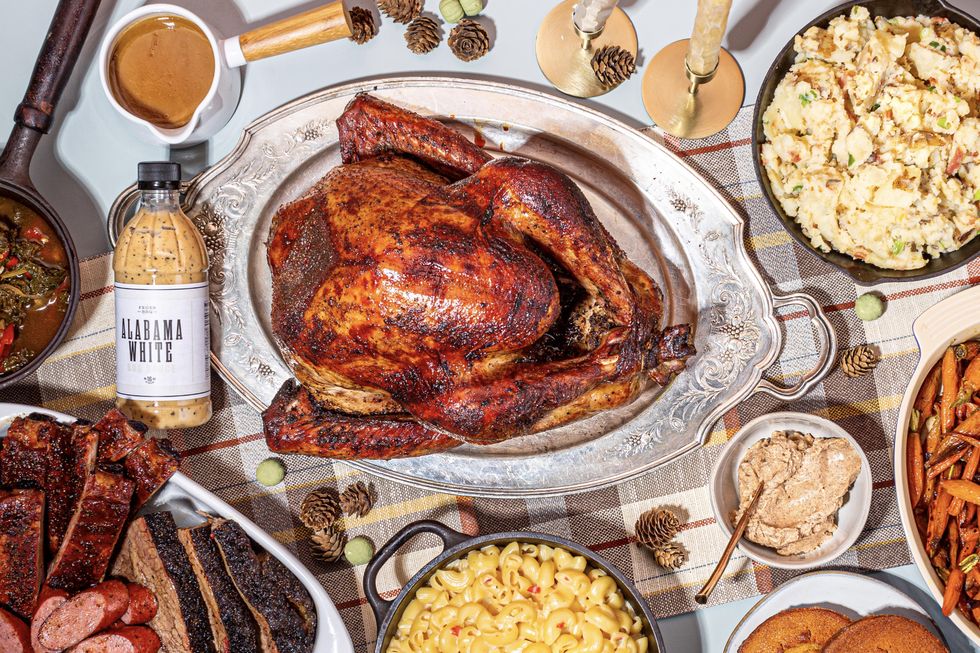 Picture of a roast turkey on a platter surrounded by plates of sausage and brisket, mac and cheese, potatoes, carrots and a jar of Alabama White sauce