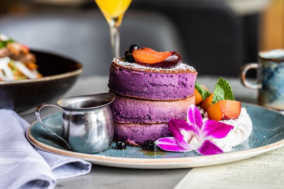 Picture of three thick stacked Japanese ube pancakes that are purple in color, on a plate with a silver pitcher of syrup and an orchid.