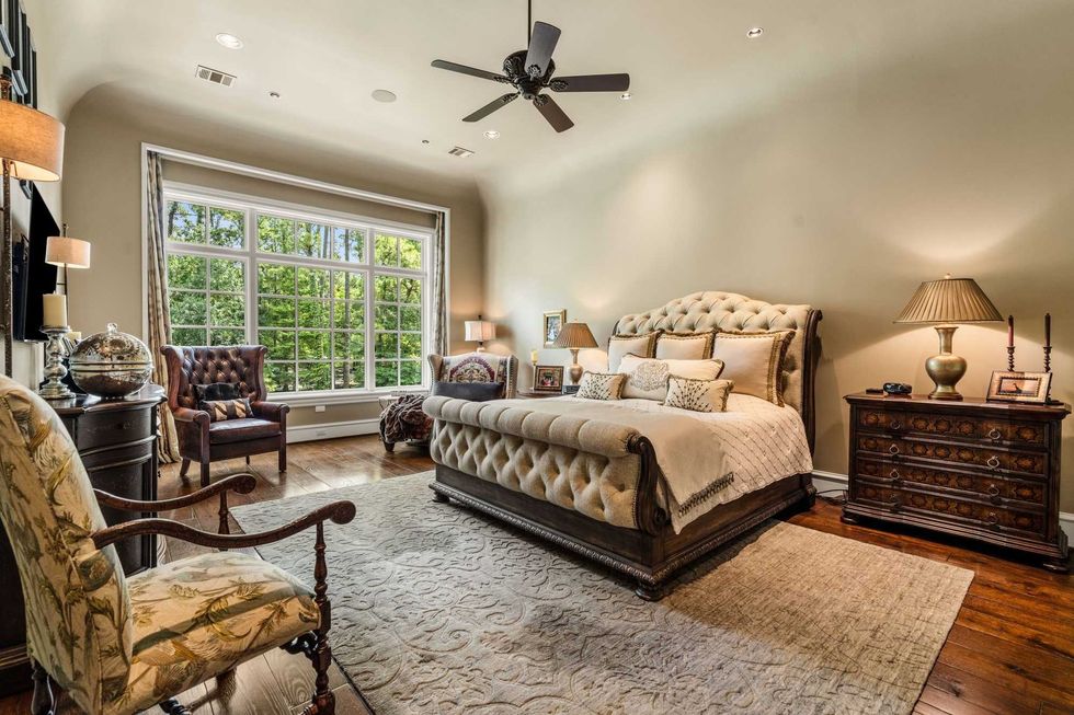The primary suite of 50 Patti Lynn Lane shows hardwood floors, a king-size bed and oversize windows overlooking wooded views