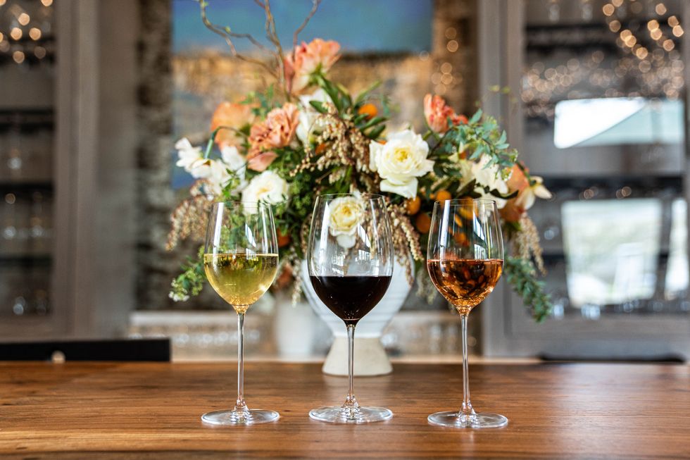 Three glasses of wine, a white, a red and a rose, on a counter with a huge vase of flowers behind them.