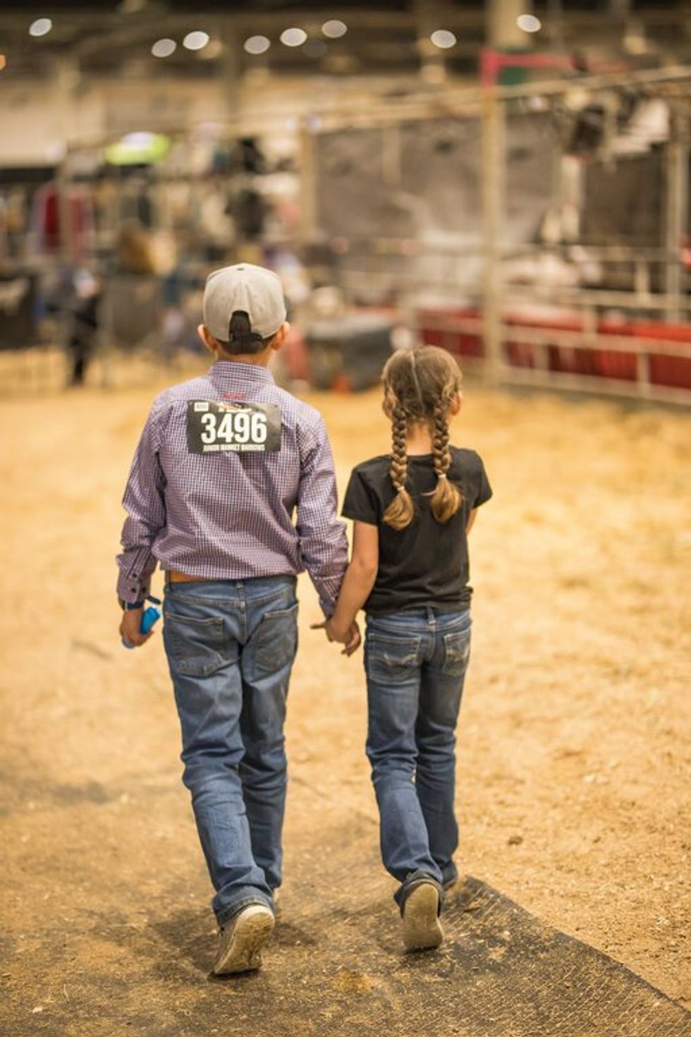 Two children, a boy and girl, with their backs to the camera, walking along a dirt-lined arena at the rodeo